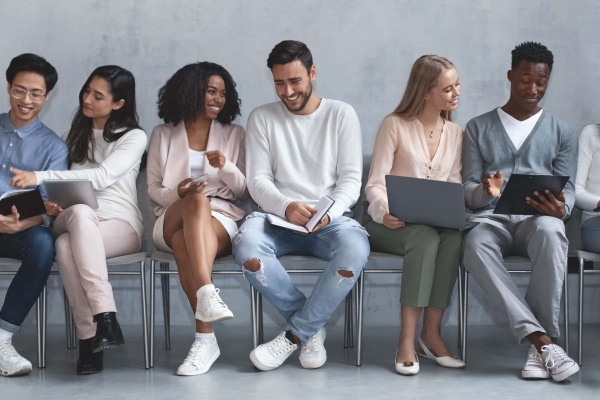 Seven casually dressed, jovial young people of different genders and ethnicities sit in a row chatting and laughing. The background is soft grey.