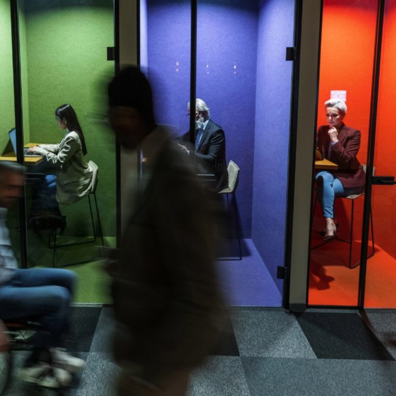 Four colleagues work individually inside multi-coloured booths while two silhouetted colleagues - one walking, one using a wheelchair - pass outside.
