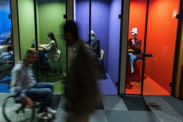 Four colleagues work individually inside multi-coloured booths while two silhouetted colleagues - one walking, one using a wheelchair - pass outside.