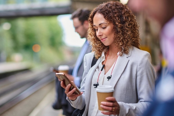 A white woman with brown hair and grey blazer holds a coffee cup in one hand while looking at a mobile in the other hands, while stood on a railway station platform.