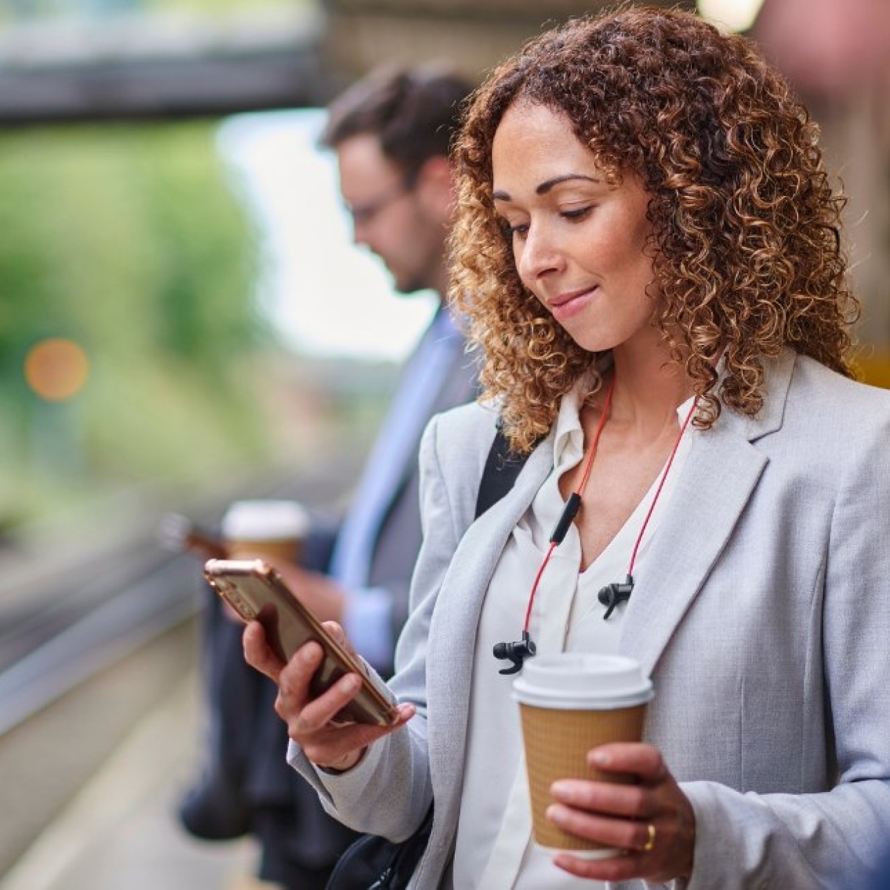 A white woman with brown hair and grey blazer holds a coffee cup in one hand while looking at a mobile in the other hands, while stood on a railway station platform.
