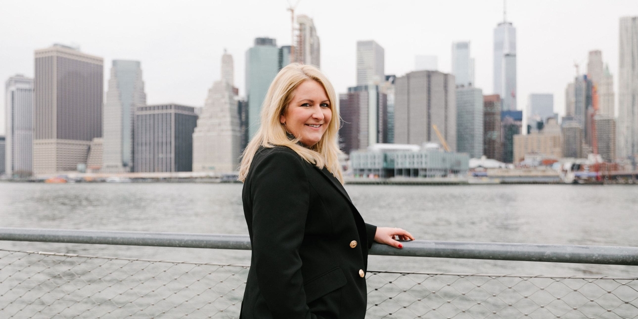Laura Sutherland stood next to the river with the New York skyline in the background on a grey day. Laura is a woman with blonde hair who wears black clothing