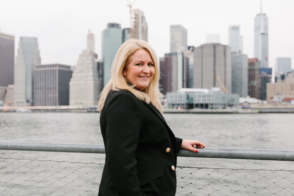 Laura Sutherland stood next to the river with the New York skyline in the background on a grey day. Laura is a woman with blonde hair who wears black clothing