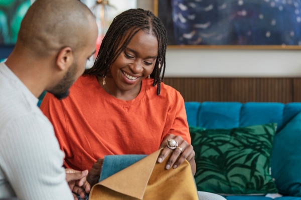 A male and female black couple sat in a furniture shop choosing sofa fabrics. They are both black; the man wears a grey top and the woman wears an orange top.