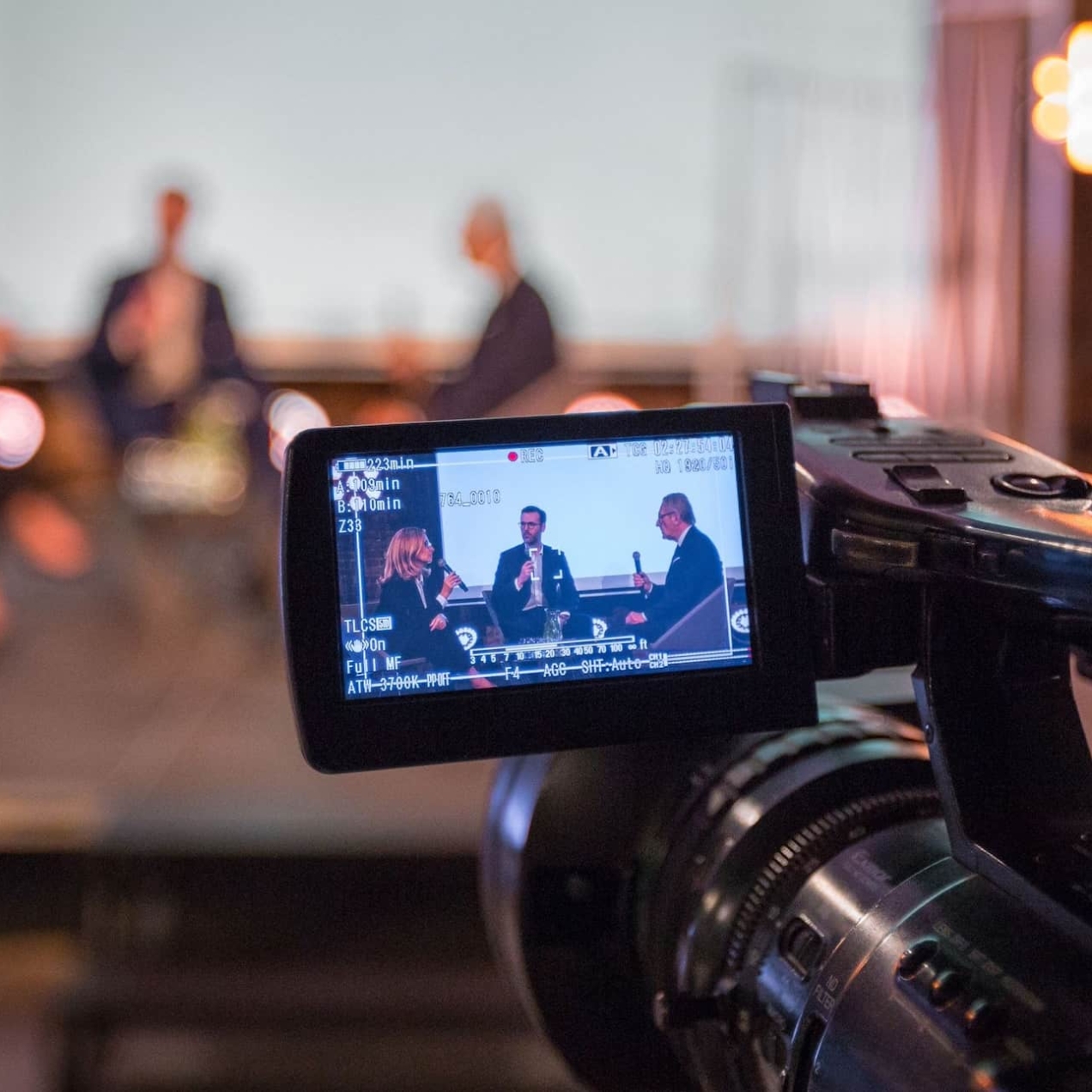 A close up of the view screen of a video camera which shows two men and one woman sat on stage speaking into microphones. The picture is replicated in the background albeit blurred