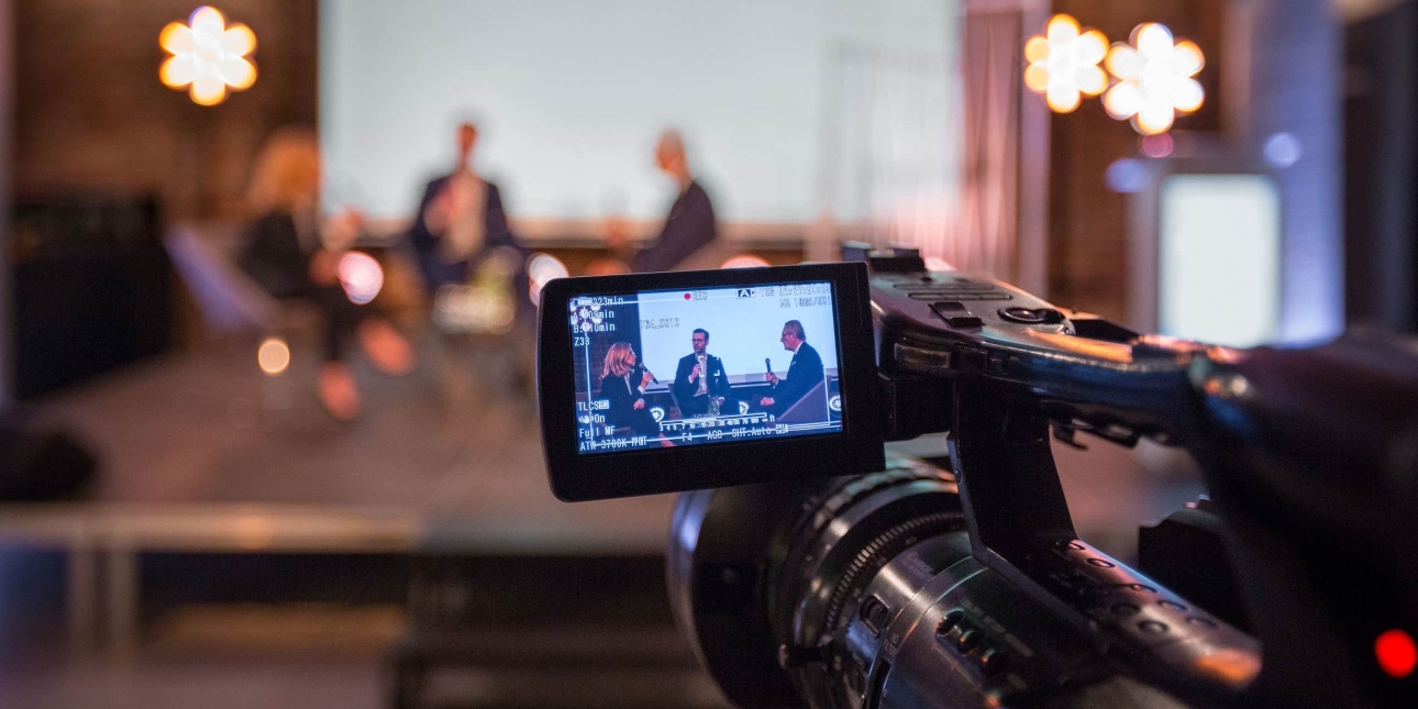 A close up of the view screen of a video camera which shows two men and one woman sat on stage speaking into microphones. The picture is replicated in the background albeit blurred