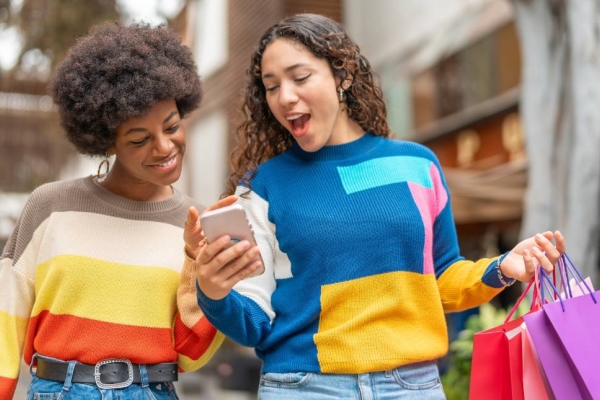 Two young women in brightly coloured jumpers smile while looking at the a mobile phone that one of them is holding.