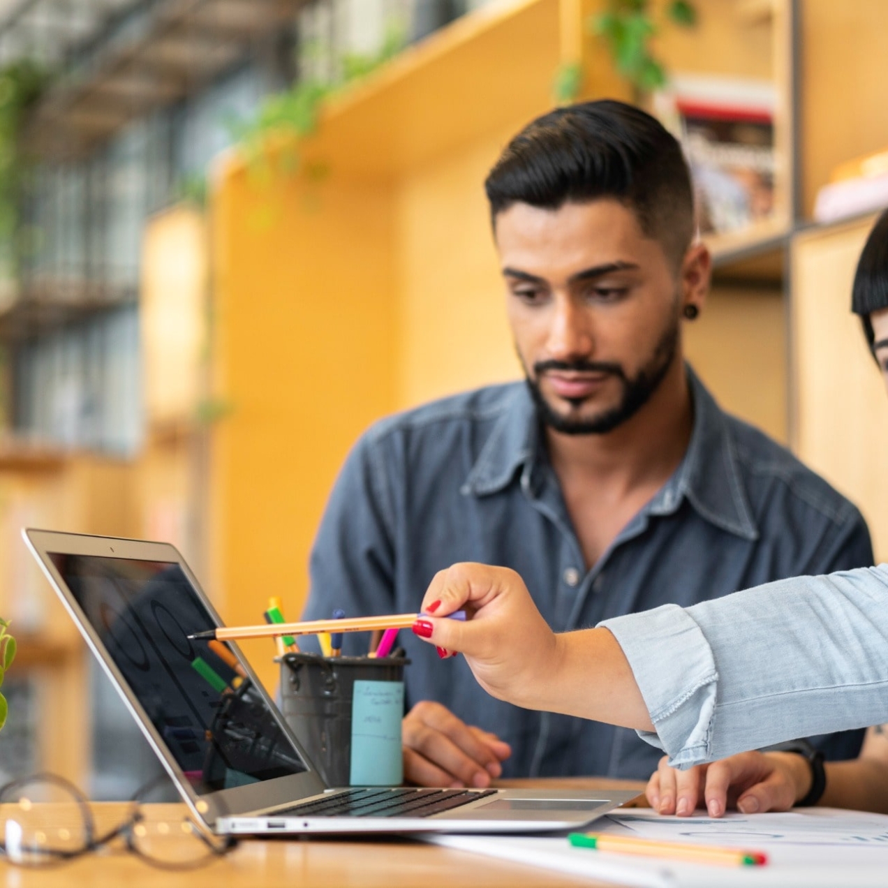 Three young, smiling colleagues gather around a laptop: an Asian man with dark hair and groomed stubble and blue shirt; an Asian woman with dark hair scratches her face; and an Asian woman with dark hair and blue shirt points a pen at a laptop screen