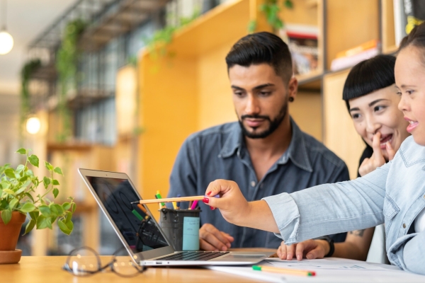 Three young, smiling colleagues gather around a laptop: an Asian man with dark hair and groomed stubble and blue shirt; an Asian woman with dark hair scratches her face; and an Asian woman with dark hair and blue shirt points a pen at a laptop screen