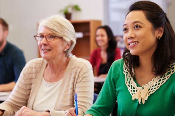 Two women sat at a desk with open notebooks in a classroom. The spectacle-wearing woman on the left is older, white, with short grey hair. The younger woman on the right is Asian, with shoulder length dark hair and wears a green top. Out of focus beh