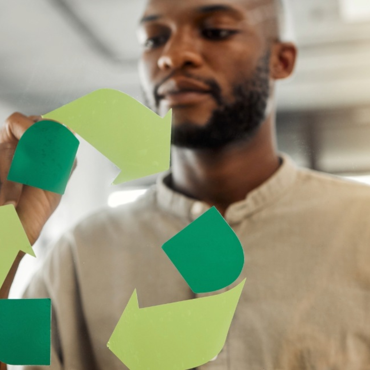 A Black man in an office, wearing a beige shirt, fixes a green recycling icon to a pane of glass.