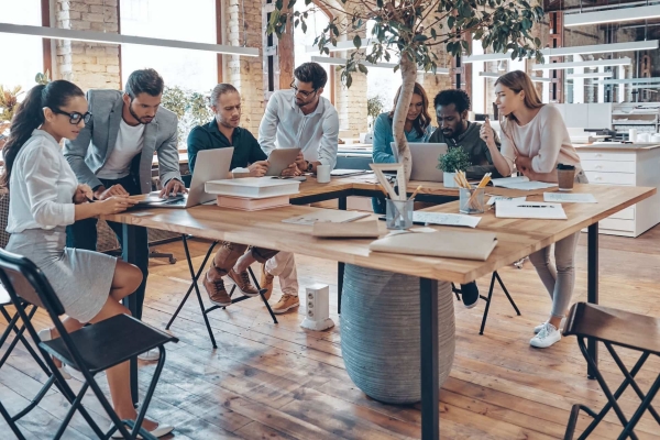 A group of six colleagues of different gender and ethnicities huddled around a table. They are concentrating on paper work and computers. There is a large plant in the middle of the desk.