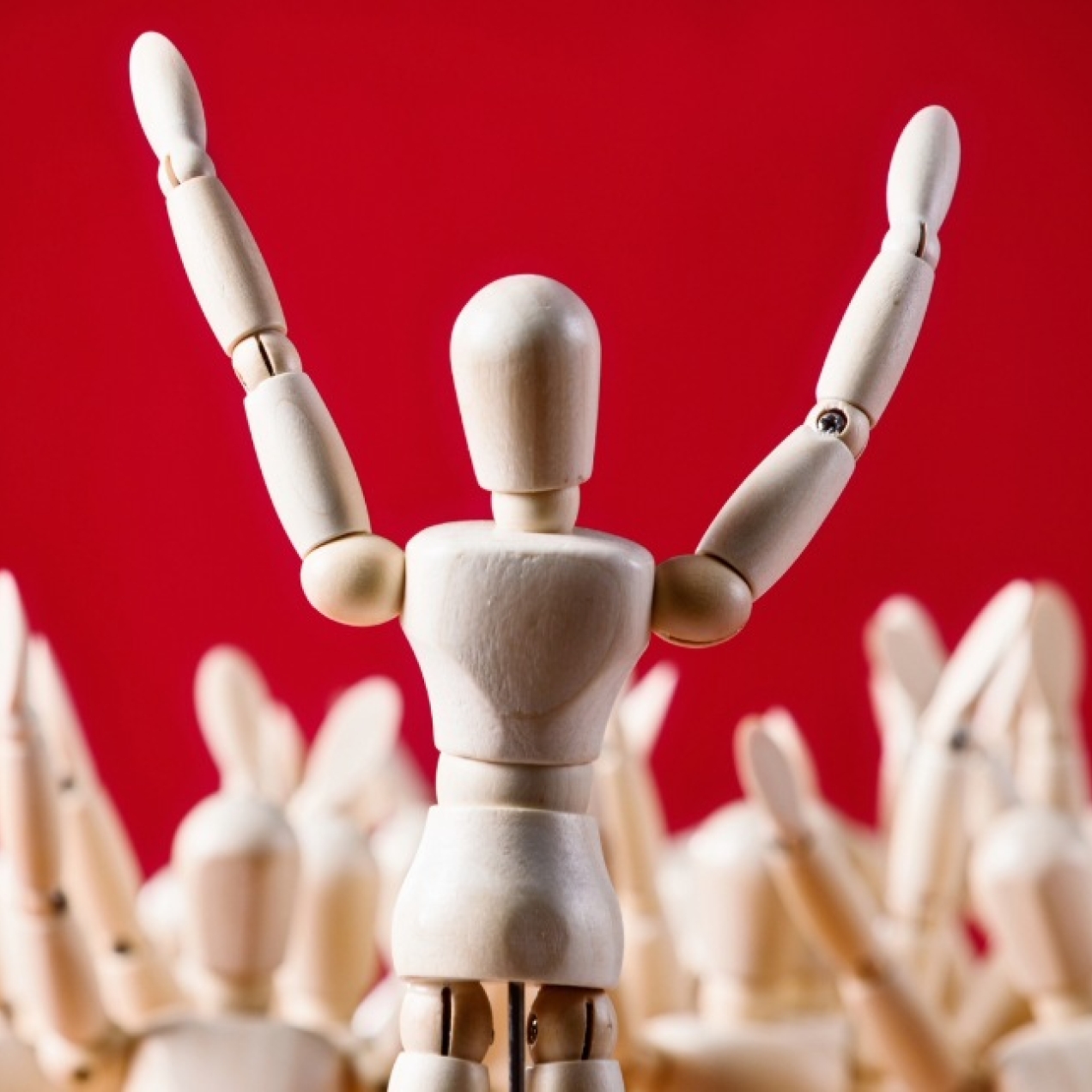 On a red background a wooden artist's mannequin stands with its arms raised addressing a crowd of other manequins.