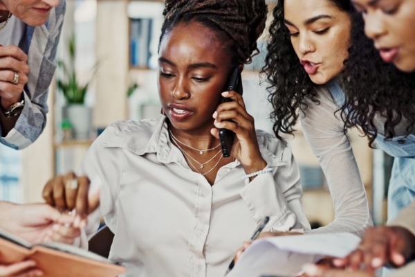 A Black business woman is sat multi-tasking as four colleagues of different genders and ethnicities try to get her attention with notebooks and documents to sign.