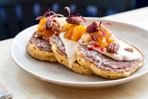 Three pancakes on a white plate on a table, piled with fruit and nuts