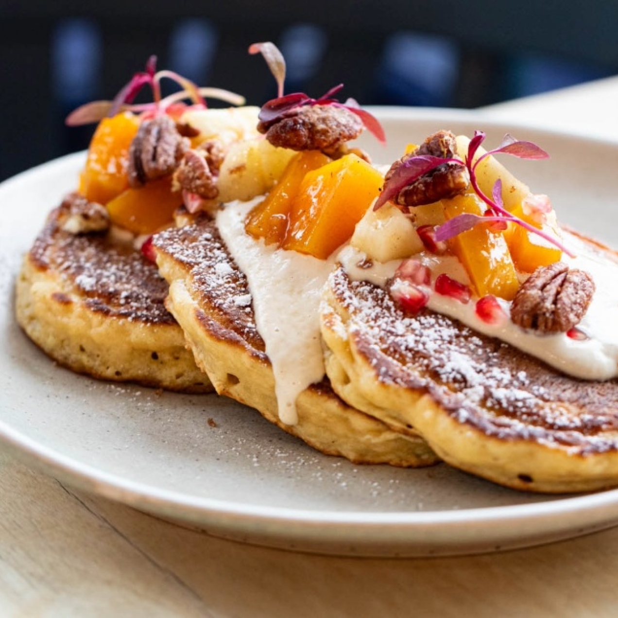 Three pancakes on a white plate on a table, piled with fruit and nuts