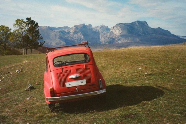 The rear of a small red vintage car parked on grass with trees and mountains in the background