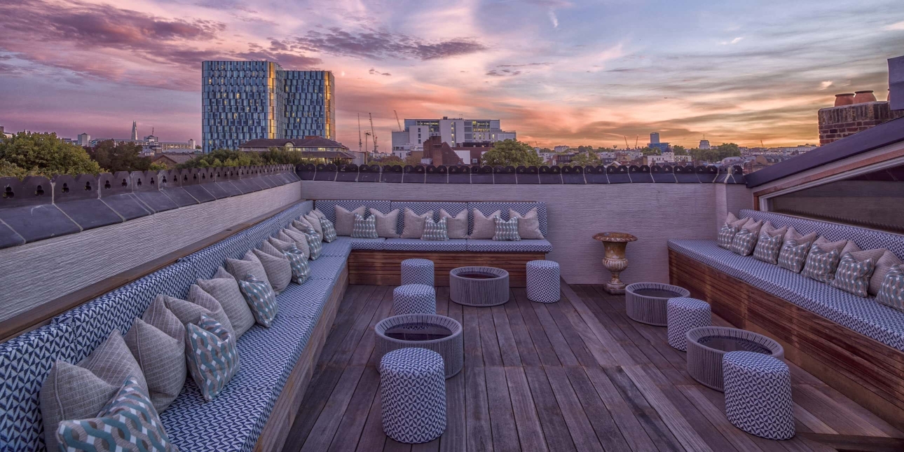 The blue styled roof terrace at The Cross London, featuring blue and white soft seating around the perimeter. The Kings Cross skyline of towers is in the background against a red-blue sky