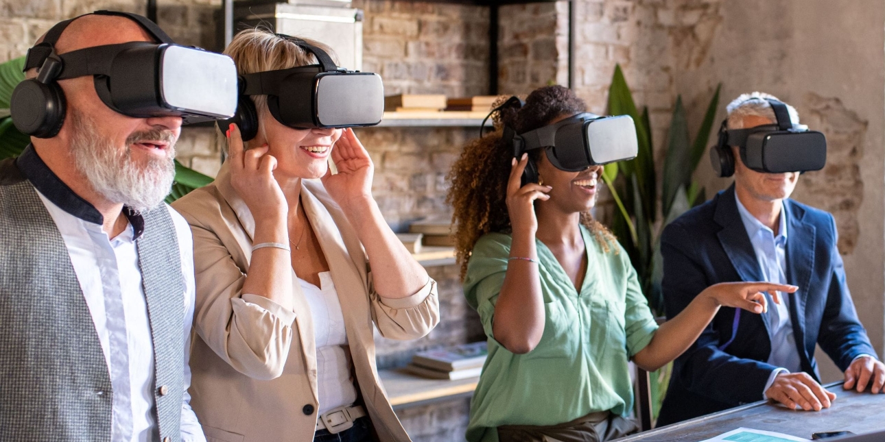 Four colleagues of different genders and ethnicities sit at a desk while wearing virtual reality headsets