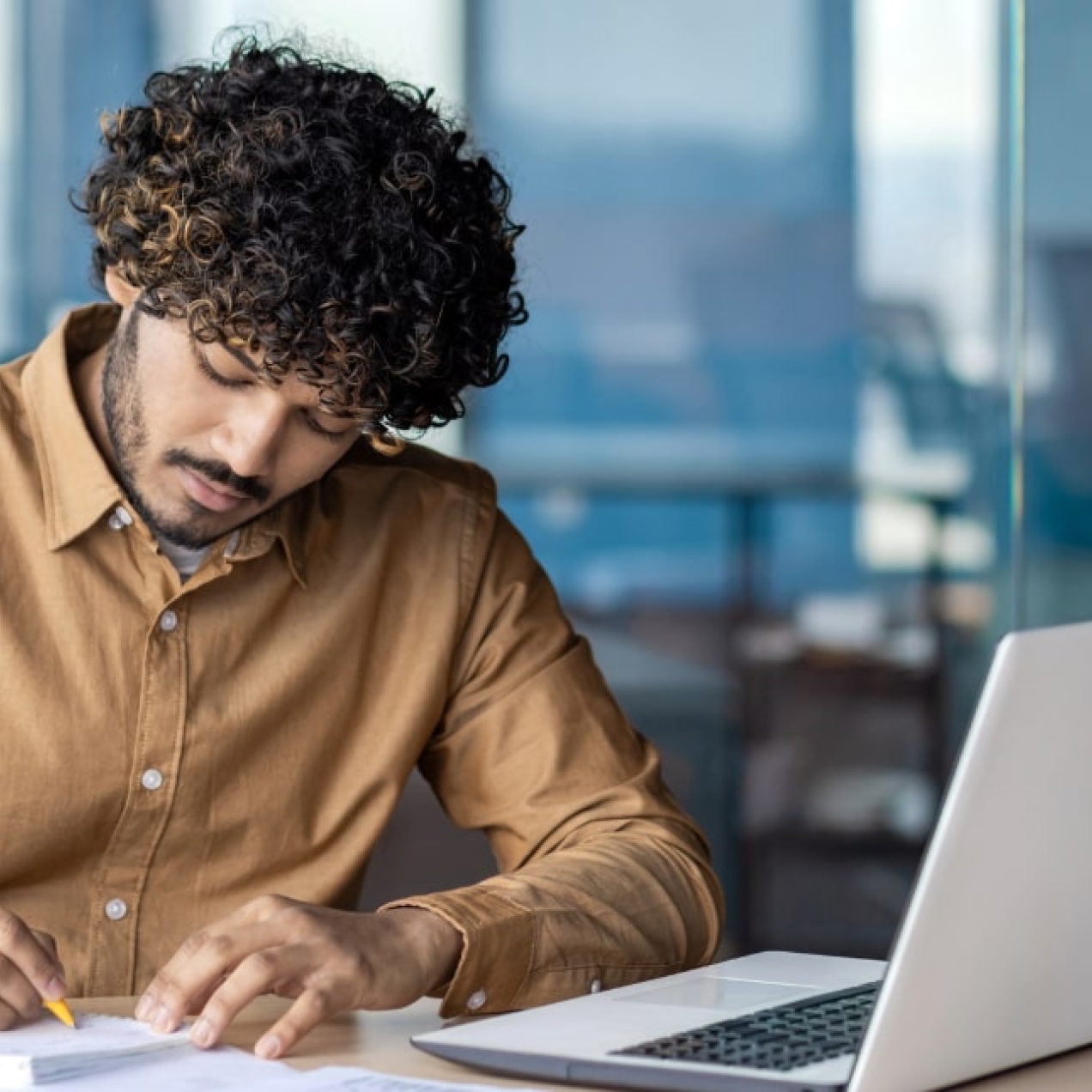 An Asian man with curly dark hair, who is wearing a mustard coloured shirt, writes on a notepad. He is in an office and there is a laptop open in front of him.