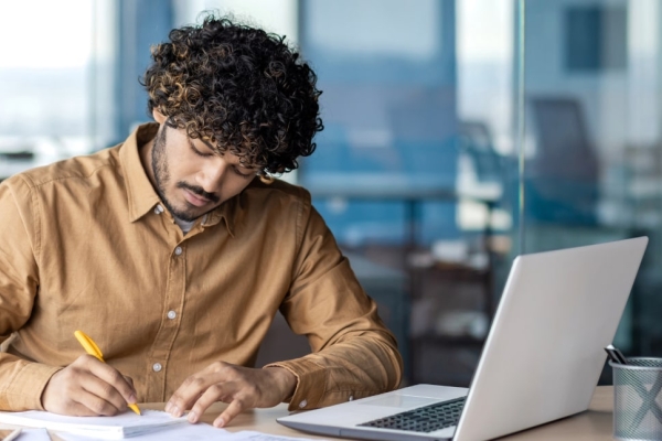 An Asian man with curly dark hair, who is wearing a mustard coloured shirt, writes on a notepad. He is in an office and there is a laptop open in front of him.