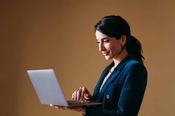 Against a brown background a business woman with dark hair and dressed in blue holds a laptop in her hand.