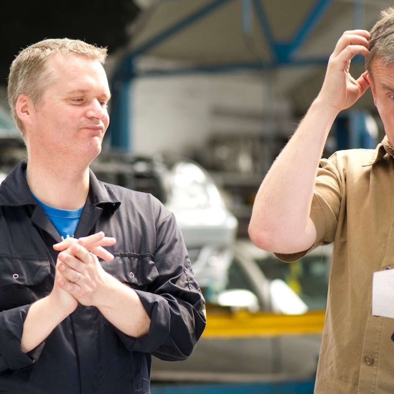 A white male mechanic rubs his hands in glee with a smile on his face. To his left a customer, who is also a white man, holds a bill in his left, scratches his head with this right hand and screws his face up in disbelief