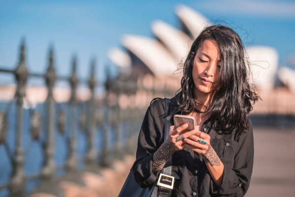 An Asian woman with long dark hair looks down at her mobile phone. The white sails of the Sydney Opera House are in the background against a blue sky.
