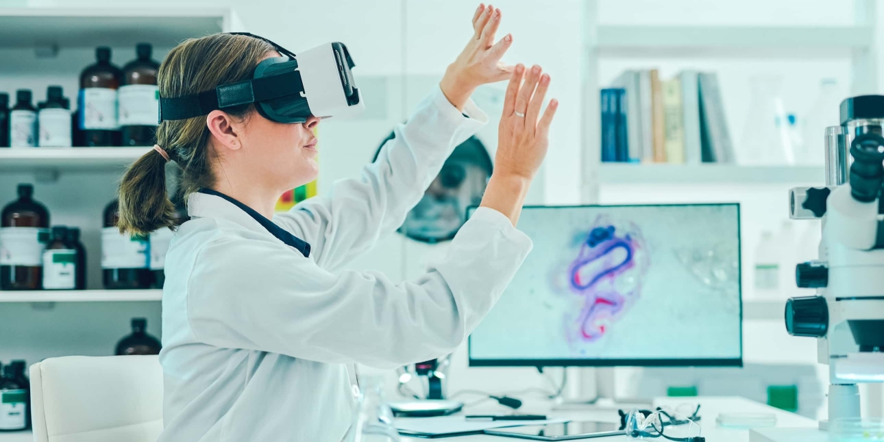 A woman scientist in a white lab coat wears a virtual reality headset inside her lab. She is waving her arms in front of her. There is technical equipment on the desk and large jars behind her