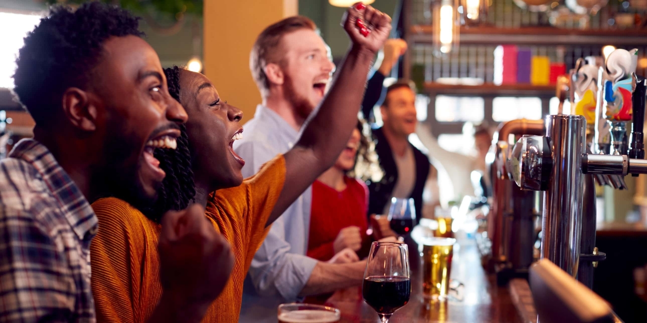Stood at the bar of a pub, a Black man, Black woman two white men and a white woman clench their fists in a celebratory gesture while looking towards a screen which out of shot