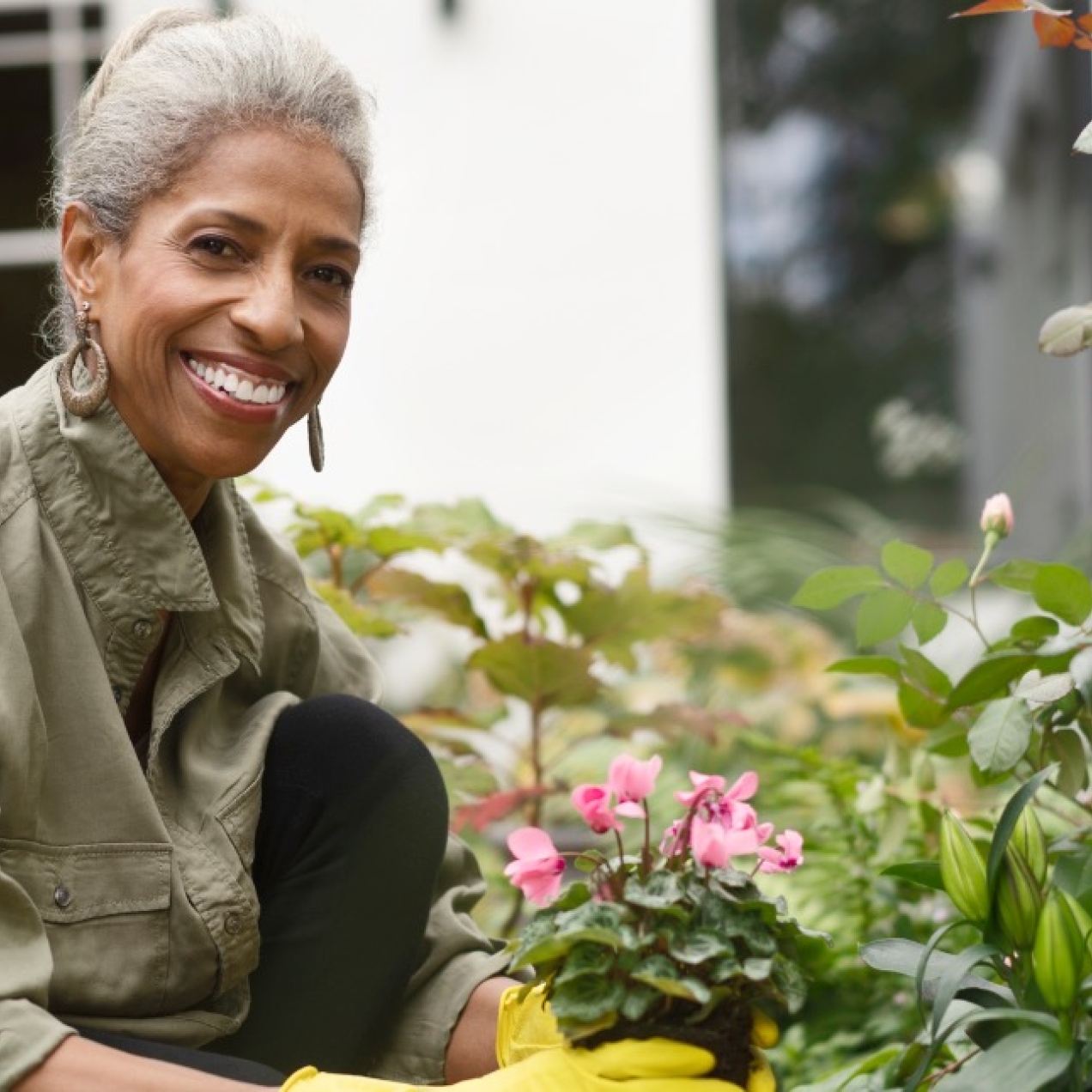 A smiling, older Black woman with grey hair, hooped earrings and khaki jacket, kneels down in her garden. Wearing yellow gloves, she is planting a pink flowering plant. Around her are green bushes a wooden bench and a house