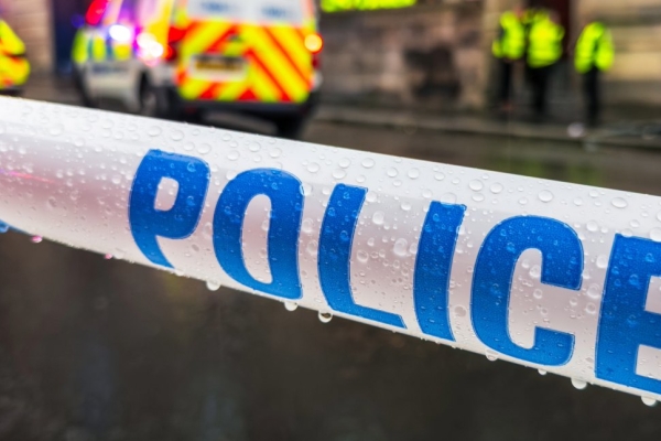Blue and white striped tape with the word police written on it. In the background are several out-of-focus police officers wearing yellow hi-vis and two police vehicles