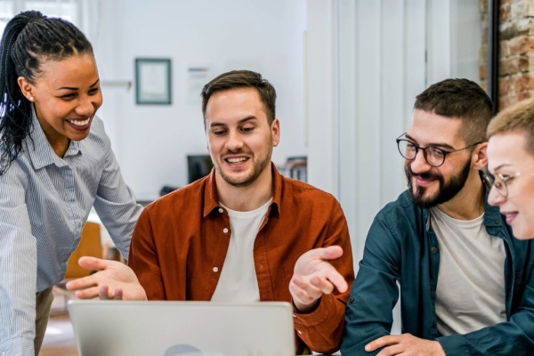 A group of male and female colleagues in a modern office stood and sat smiling around a laptop.