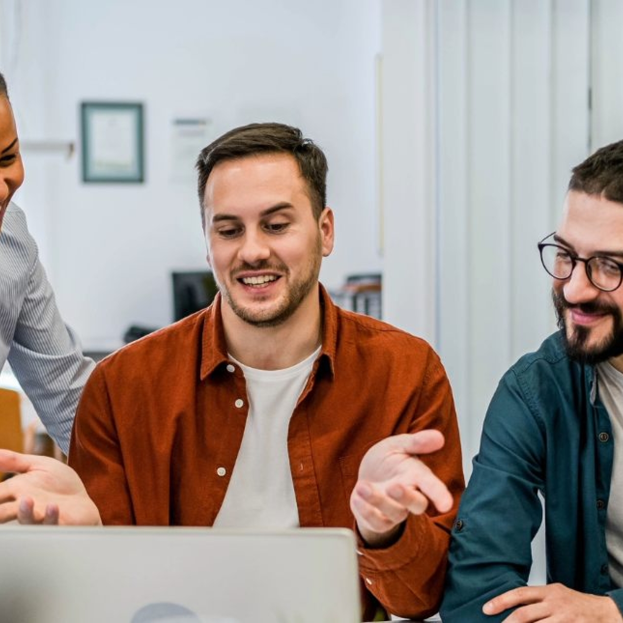 A group of male and female colleagues in a modern office stood and sat smiling around a laptop.