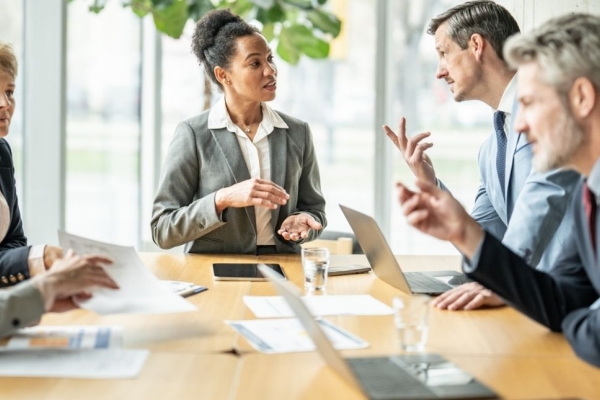 Five board members of different genders and ethnicities engage in a serious discussion around a table in an office.