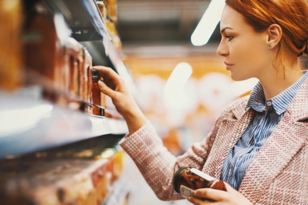 A white woman with red hair, who is wearing a plaid jacket, holds a jar of sun-dried tomatoes while browsing supermarket shelves.