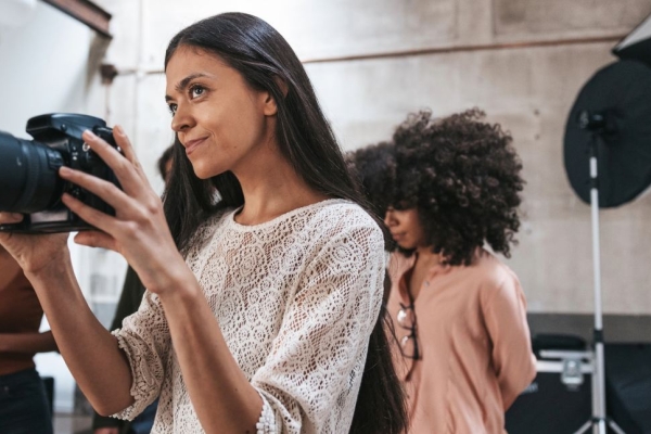 An Asian female photographer wearing a white laced top points a long lens camera in her hands while other women stand behind her. Also in background are photographic lights and a cement wall.
