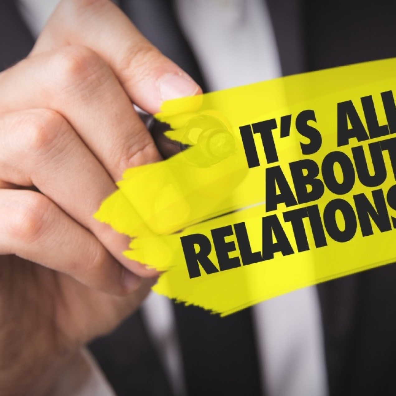 A close up of the hand of a white male wearing a black suit. He is holding a pen to a transparent board. Superimposed in black writing on a yellow background are the words: it's all about relationships.