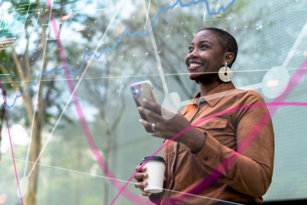 A smiling Black woman holds a phone in one hand and a coffee cup in the other, while stood beneath trees.