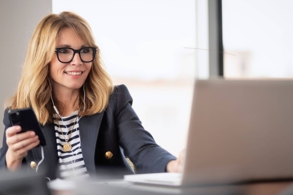 A middle-aged white woman with shoulder length blonde hair sits in front of a laptop with earphones connected to her mobile. She wears glasses and a navy jacket over a navy and white striped top