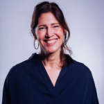 A colour portrait of Aby Hawker on a light blue background.She has long dark hair and wears a dark v-neck top.