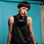 A colour photograph of Alina Smyshliak-Boroda stood in front of a turquoise background. Alina is a white woman with short dark hair wearing a black sleeveless top.