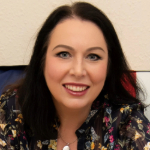 A colour portrait of Alison Gallagher-Hughes. Alison is a white woman with shoulder length dark hair who wears a patterned shirt.