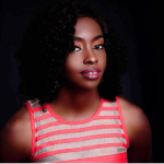 A colour portrait of Amina Maikori lookingside on at the camera. Aminia is a Black woman with shoulder length dark hair. She wears a striped pink and silver vest.
