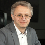 A colour photograph of Andrius Kasparas against a black background. Adrius is a white man with grey hair wearing glasses and a grey suit jacket over a white shirt.