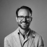 A black and white portrait of Carl Ronander smiling at the camera. Carl is a white man with short dark hair and beard, who wears glasses and an open neck shirt