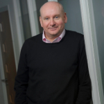 A colour photograph of Chris Lewis stood in front of a window. Chris is a white man wearing a dark jumper.