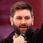 On a maroon background a colour photograph of Daniel Coole. Daniel is a white man with short dark hair and beard, wearing a dark shirt and holding a microphone in his right hand.
