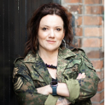 A colour portrait of Dannie-Lu Carr stood with her arms crossed against a brick wall. Dannie-Lu is a white woman with dark hair wearing a camouflage jacket.