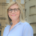A colour portrait of Dee Cowburn in front of a sandstone building. Dee is a white woman with a blond hair and a blue shirt.